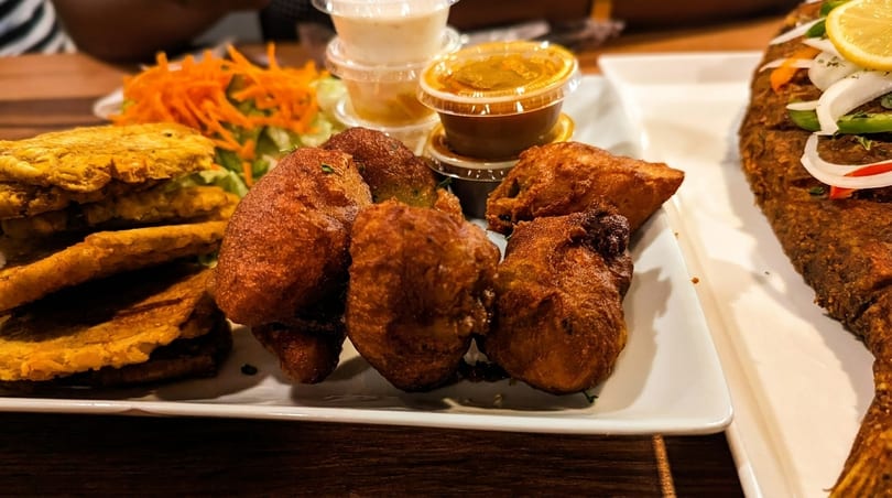 A platter of fried fish, plantains, fritters, salad, and dipping sauces on a wooden table.