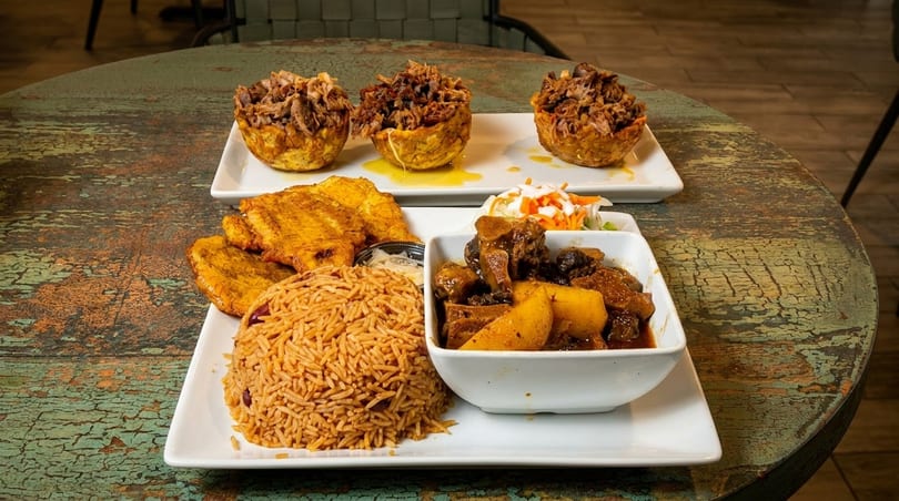 Two plates of Caribbean food on a rustic table, featuring mofongo, rice, plantains, and stew.