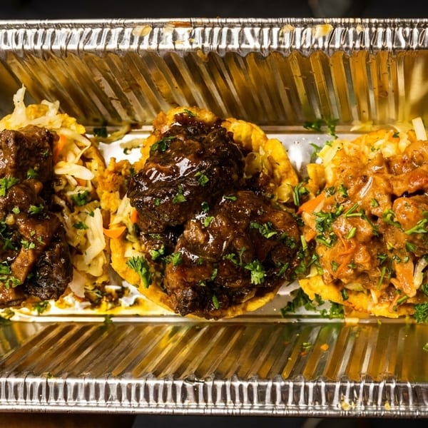 Three Caribbean tacos, two with oxtail and one with curry chicken, in a foil takeout container.
