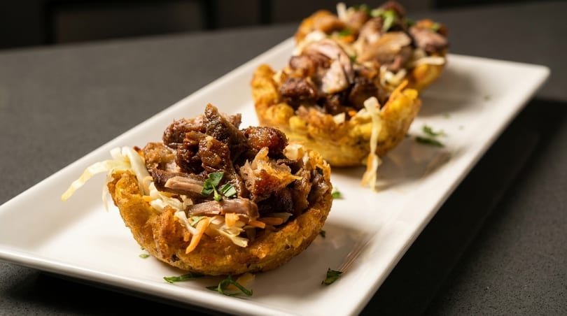 Two golden fried plantain cups filled with savory shredded meat and fresh coleslaw on a white plate.
