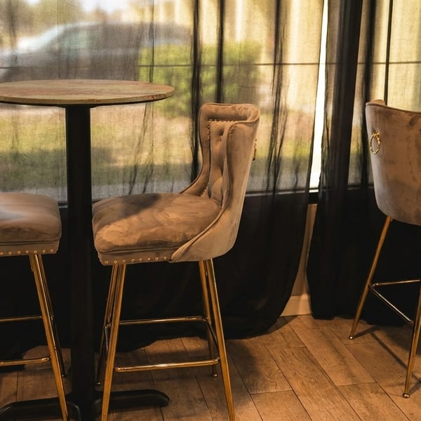 Three elegant velvet bar stools with golden legs and two round tables in a dimly lit room.