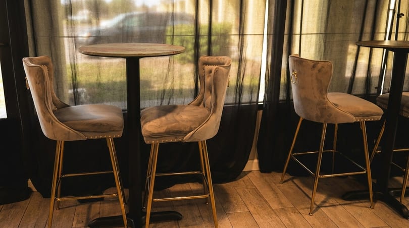 Three elegant velvet bar stools with gold legs surround two tall, round tables in a dimly lit room.