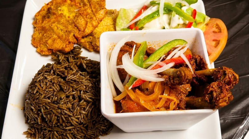 A plate of stewed meat with onions and peppers, dark rice, fried plantains, and a side salad.